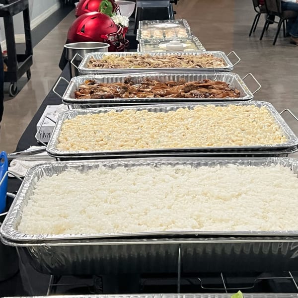 A buffet line with trays of Hawaiian food, including rice, macaroni salad, and meats.